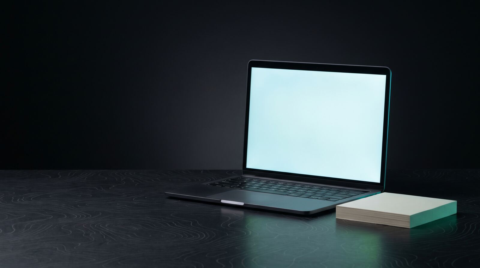Otwarty laptop z jasnym ekranem na ciemnym biurku z motywem poziomic, obok blok notesu. Claude Cowork to praca AI na plikach w folderze na komputerze.