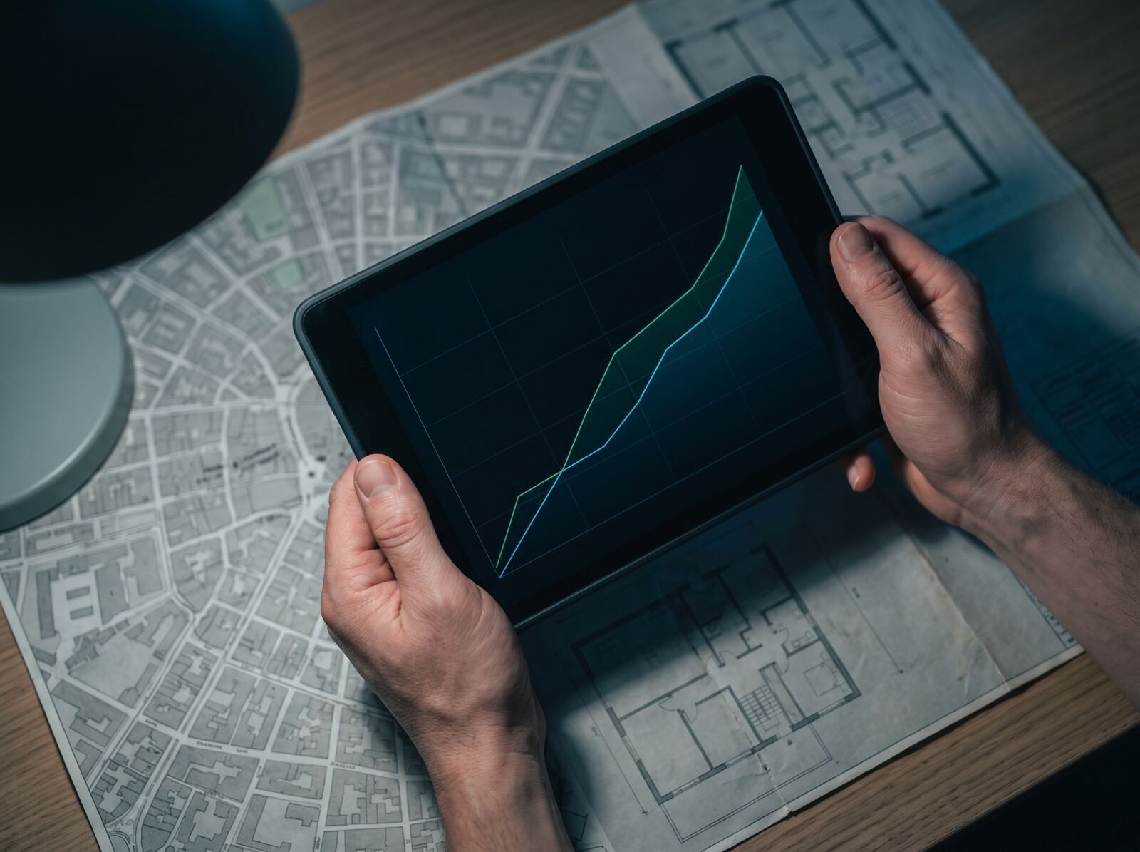 Dłonie trzymają tablet z wykresem nad mapą miasta i rzutem mieszkania. Analiza danych z rejestrów publicznych z pomocą AI.